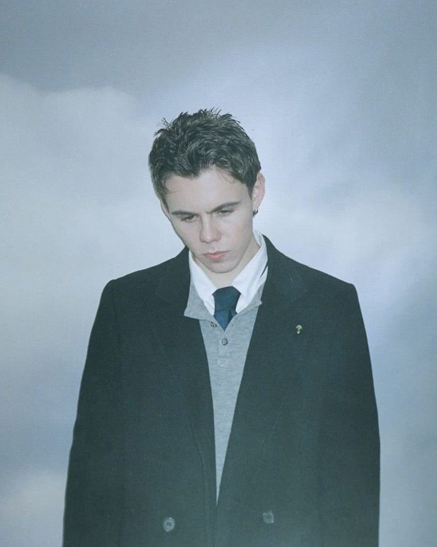 The Kid LAROI in a dark overcoat and tie looking down against a grey cloudy sky - the official A Perfect World tour image