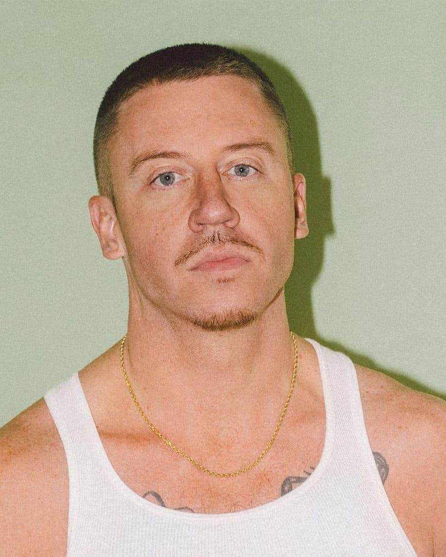 Macklemore in a white vest and gold chain against a pale green wall - the official tour image