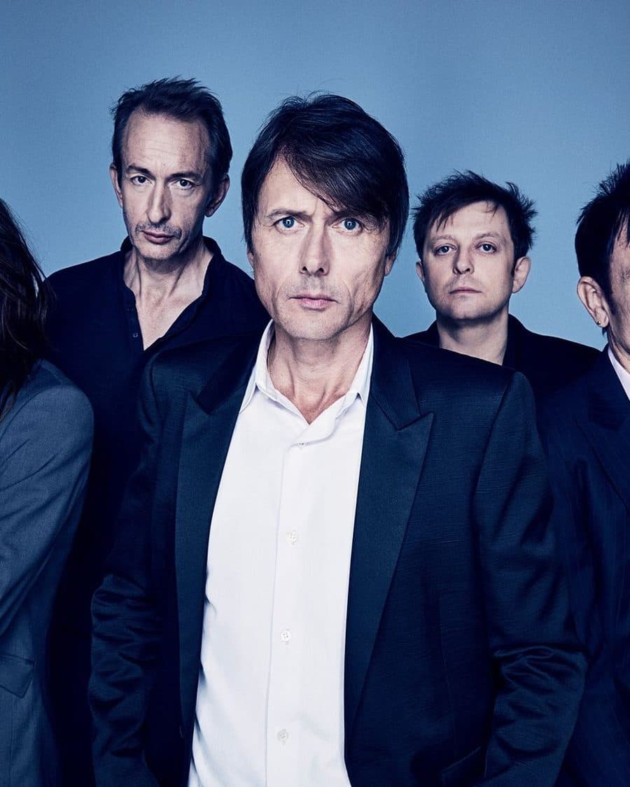 Suede's five members in dark suits against a blue studio backdrop, Brett Anderson at the front - the official listing image for the co-headline show