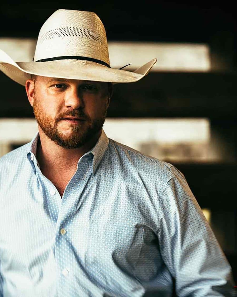 Cody Johnson in a white cowboy hat and pale blue shirt leaning against barn timbers - the official artist image