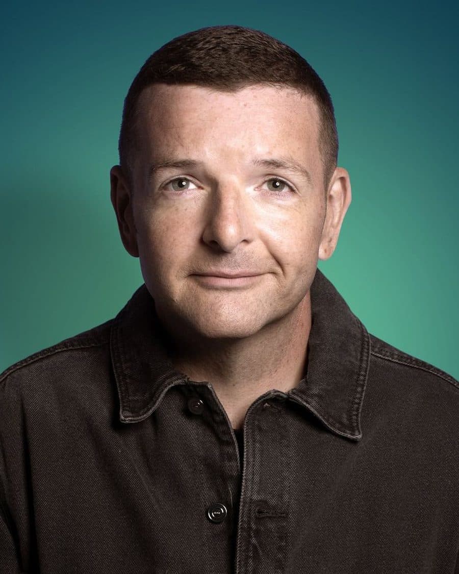 Kevin Bridges smiling in a dark denim shirt against a teal-green backdrop - the official tour portrait
