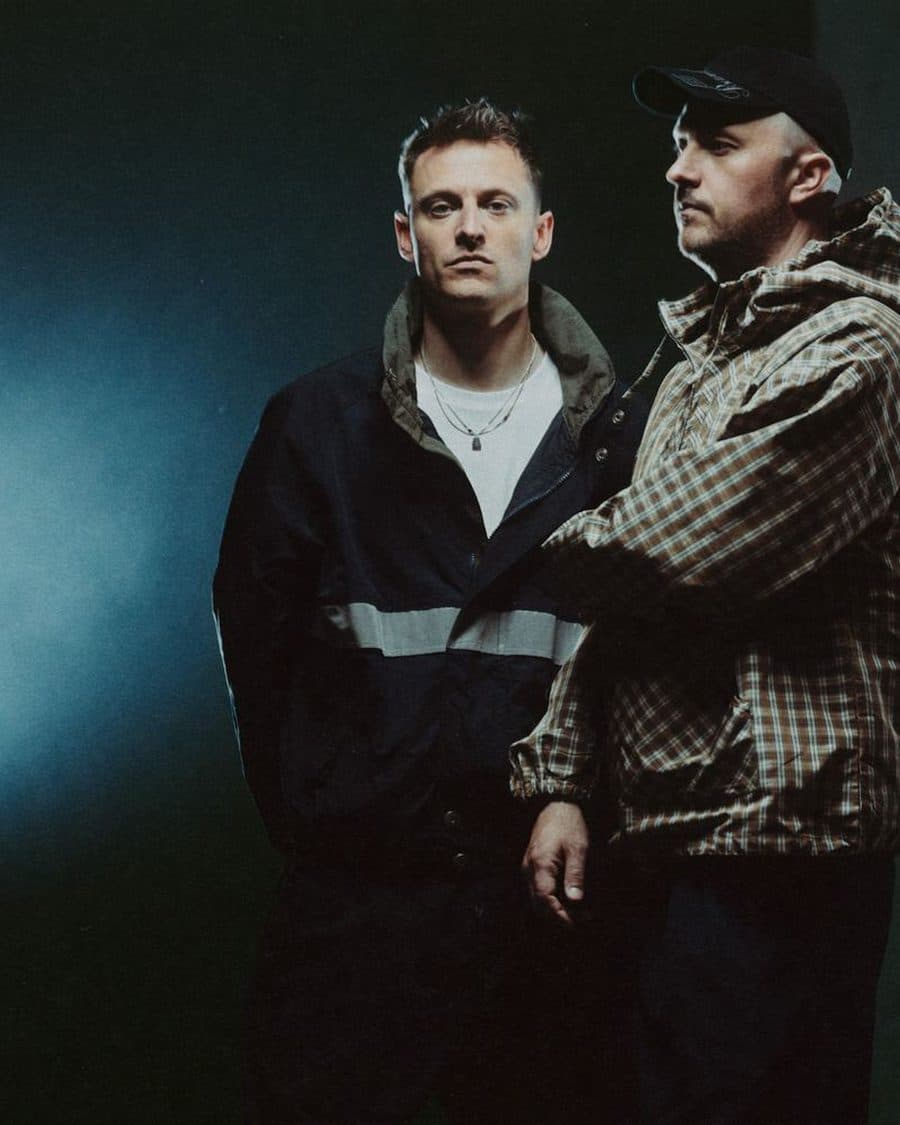 Overmono's two members in jackets and a cap lit by a pale spotlight against a dark green wall - the official duo image