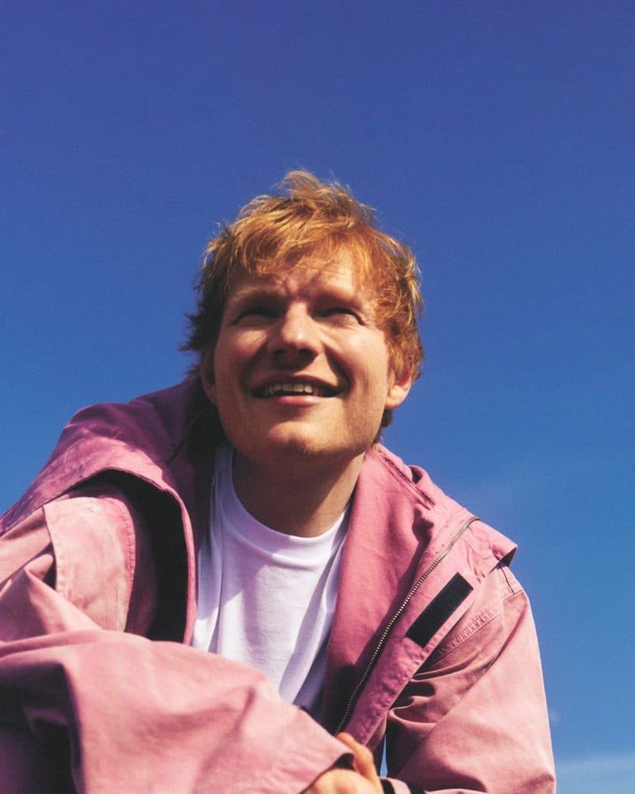 Ed Sheeran smiling in a pink jacket over a white T-shirt against a clear blue sky, shot from below - the official LOOP Tour image