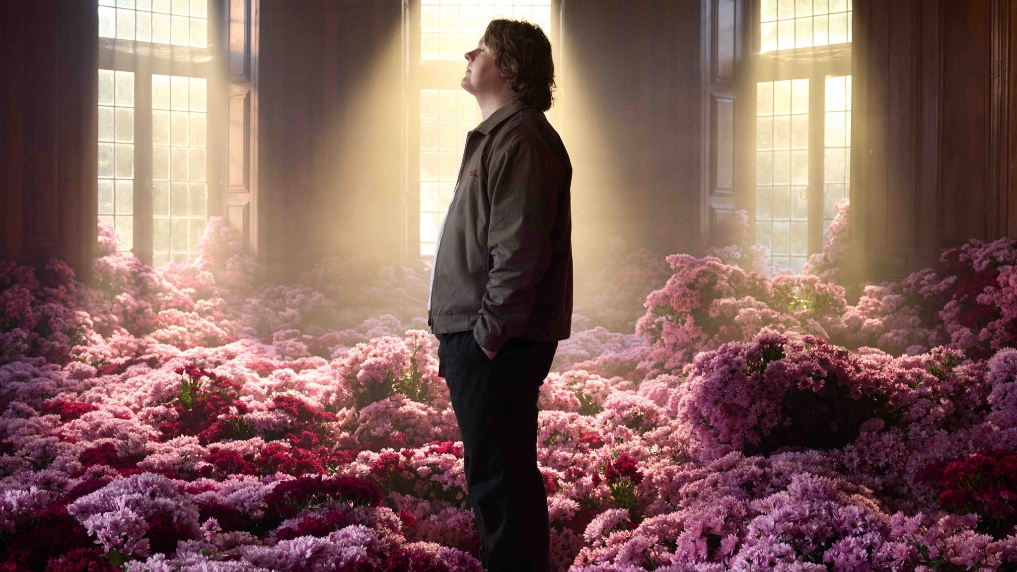 Lewis Capaldi standing in a wood-panelled room filled with pink flowers — the official tour image