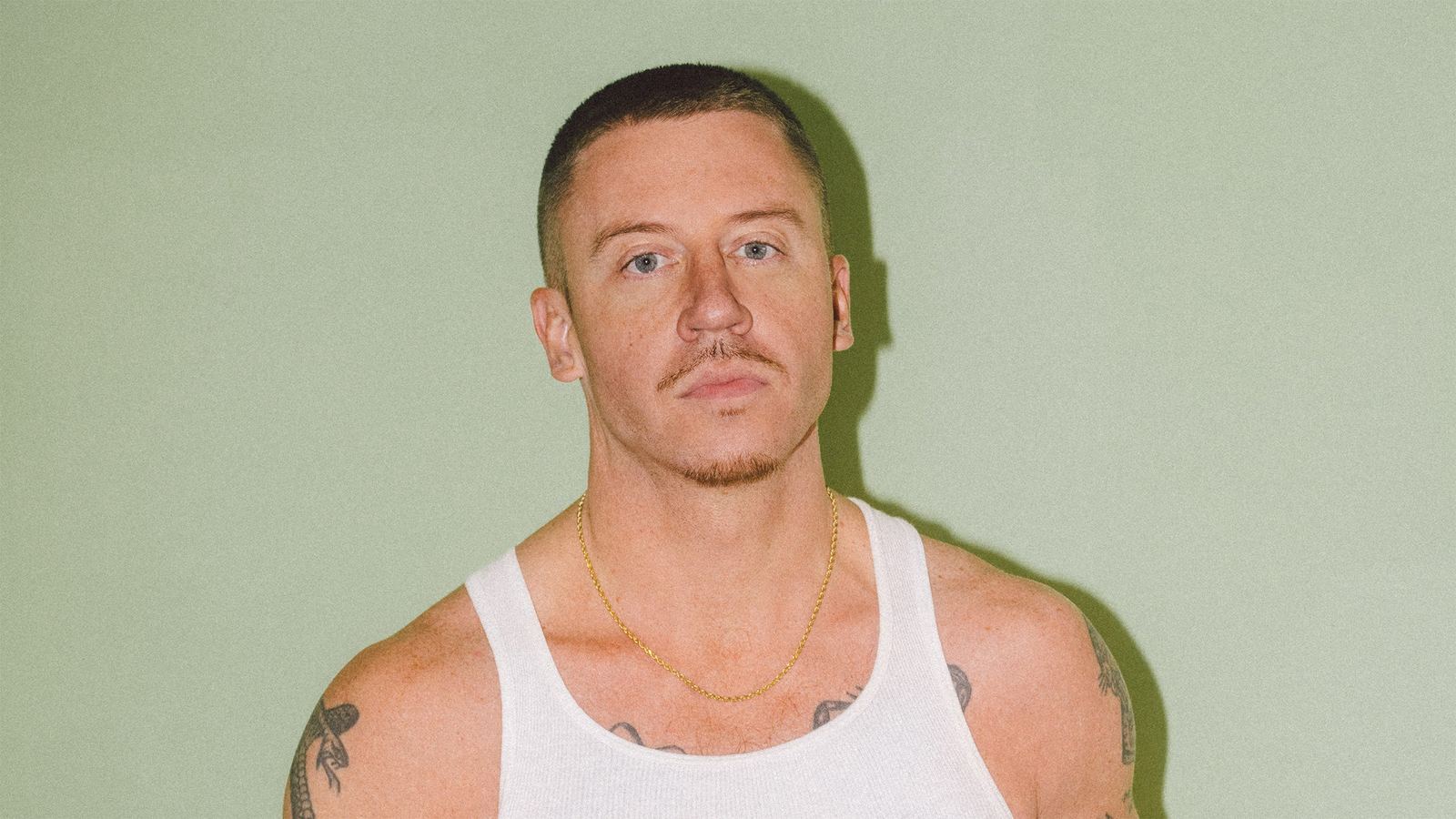 Macklemore in a white vest and gold chain against a pale green wall - the official tour image