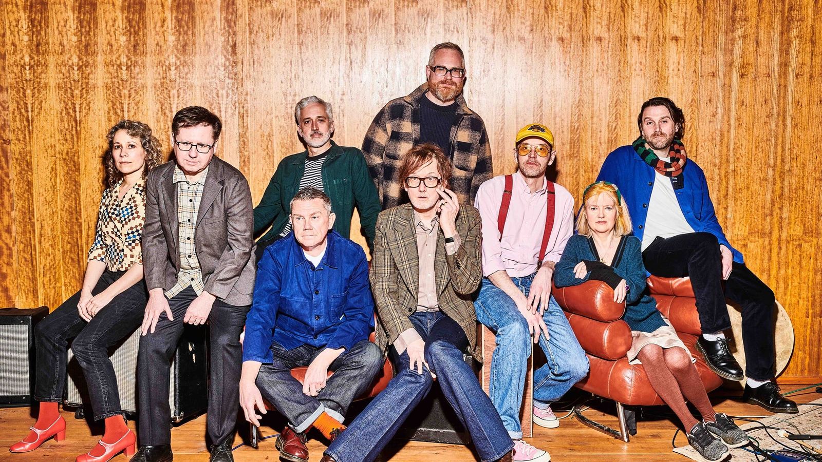 The nine members of Pulp, Jarvis Cocker front centre, seated against a wood-panelled wall — the official promo image