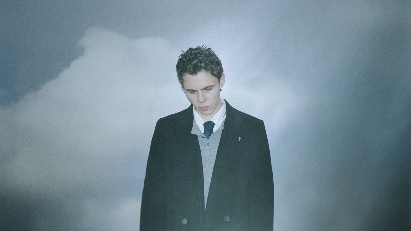 The Kid LAROI in a dark overcoat and tie looking down against a grey cloudy sky - the official A Perfect World tour image