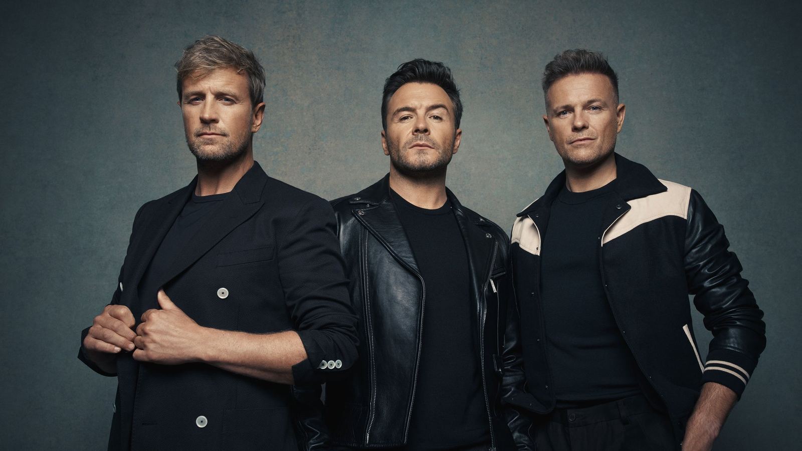 Westlife - Kian Egan, Shane Filan and Nicky Byrne in black jackets against a grey studio backdrop - the official Westlife 25 tour image